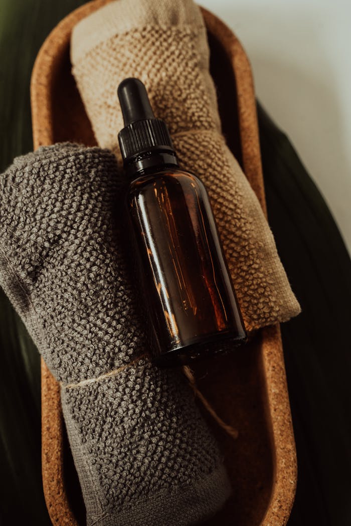 A brown glass dropper bottle on a rustic wooden tray with beige and gray towels, creating a cozy spa atmosphere.